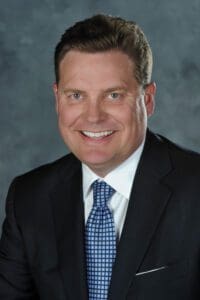 Portrait of Shane Albers, a man in a suit and blue tie smiling